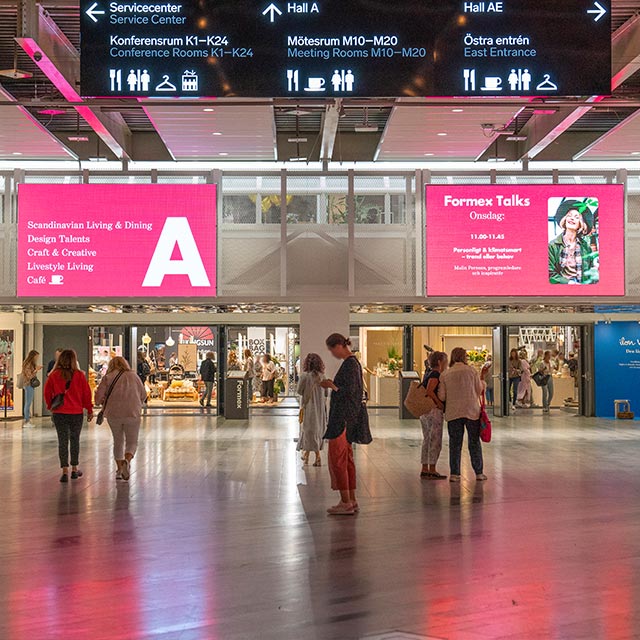på bild syns människor i rörelse framför entrén till hall A. Digitala skyltar med vägbeskrivning syns i bakgrunden.