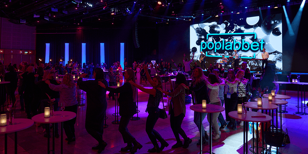 Live band performing in front of an audience, with a colourful neon-lit stage backdrop and illuminated cocktail tables during a kick-off in Victoria Hall at Stockholmsmässan